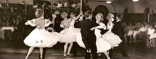 Tanzformation mit Herren in Frack und Damen in weißem Kleid tritt auf. Publikum und Band im Hintergrund.